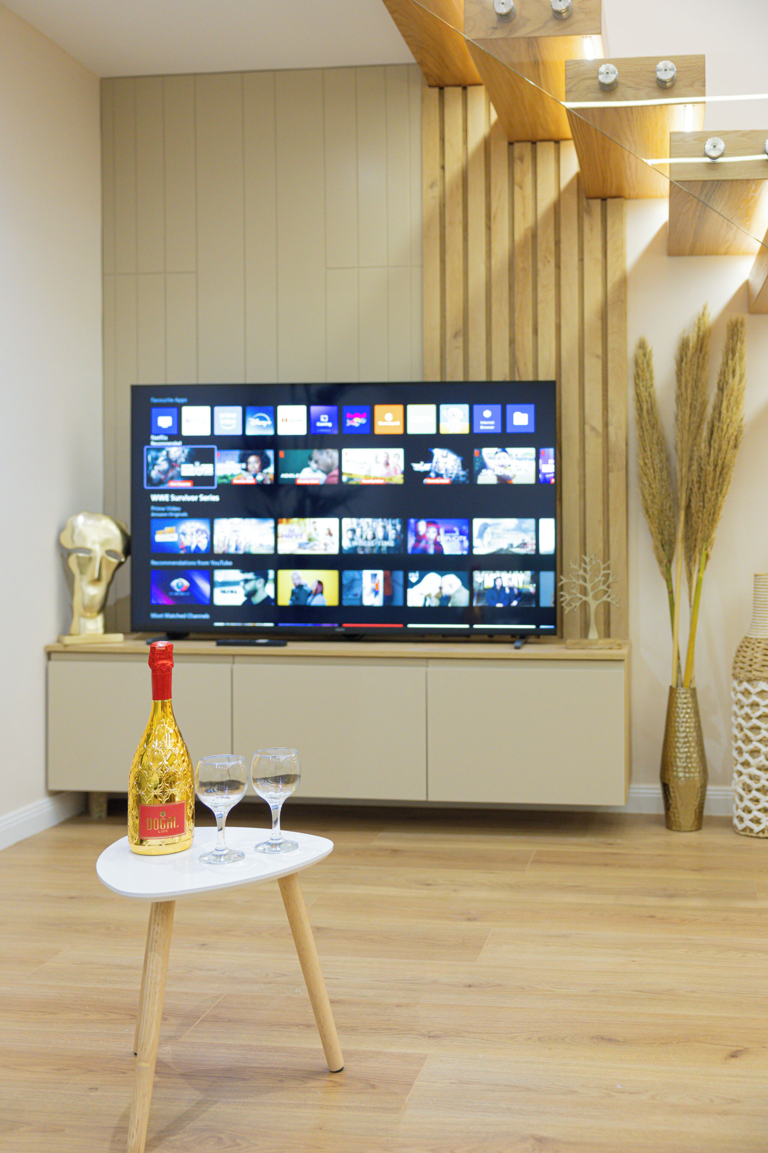 Un salon moderne avec un téléviseur à écran plat fixé au mur, une petite table sur laquelle se trouvent une bouteille et des verres, ainsi que des éléments décoratifs tels qu’un vase et des herbes hautes.