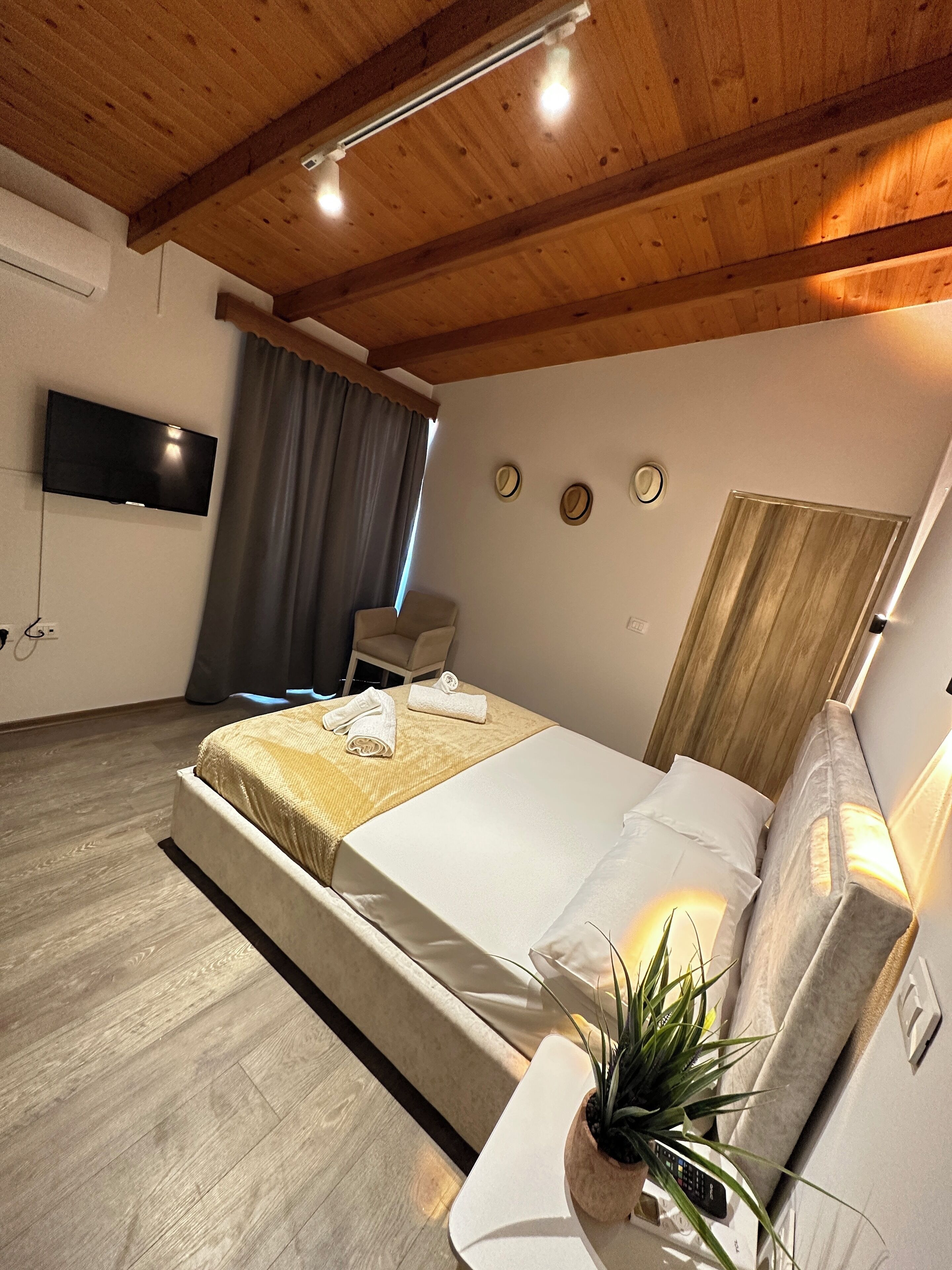 Habitación de hotel moderna con techo de madera, televisor de pantalla plana montado en la pared, cama con ropa blanca, una pequeña planta en maceta y una mesita de noche.