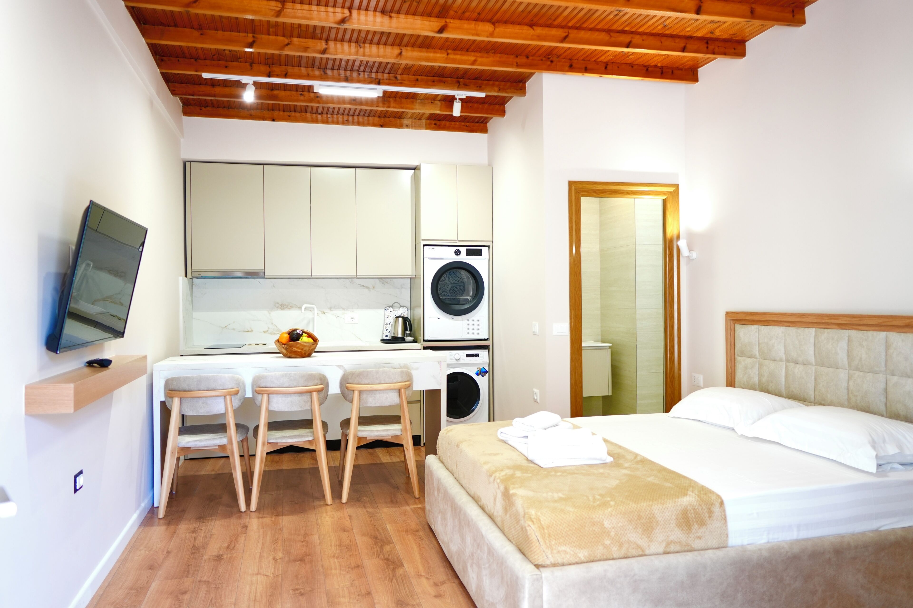 Habitación funcional con cama, escritorio y dos sillas, lavadora, televisor y techo de madera.