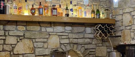 A bar area with a stone wall, wooden shelves with liquor bottles, a coffee machine, and hanging light fixtures.
