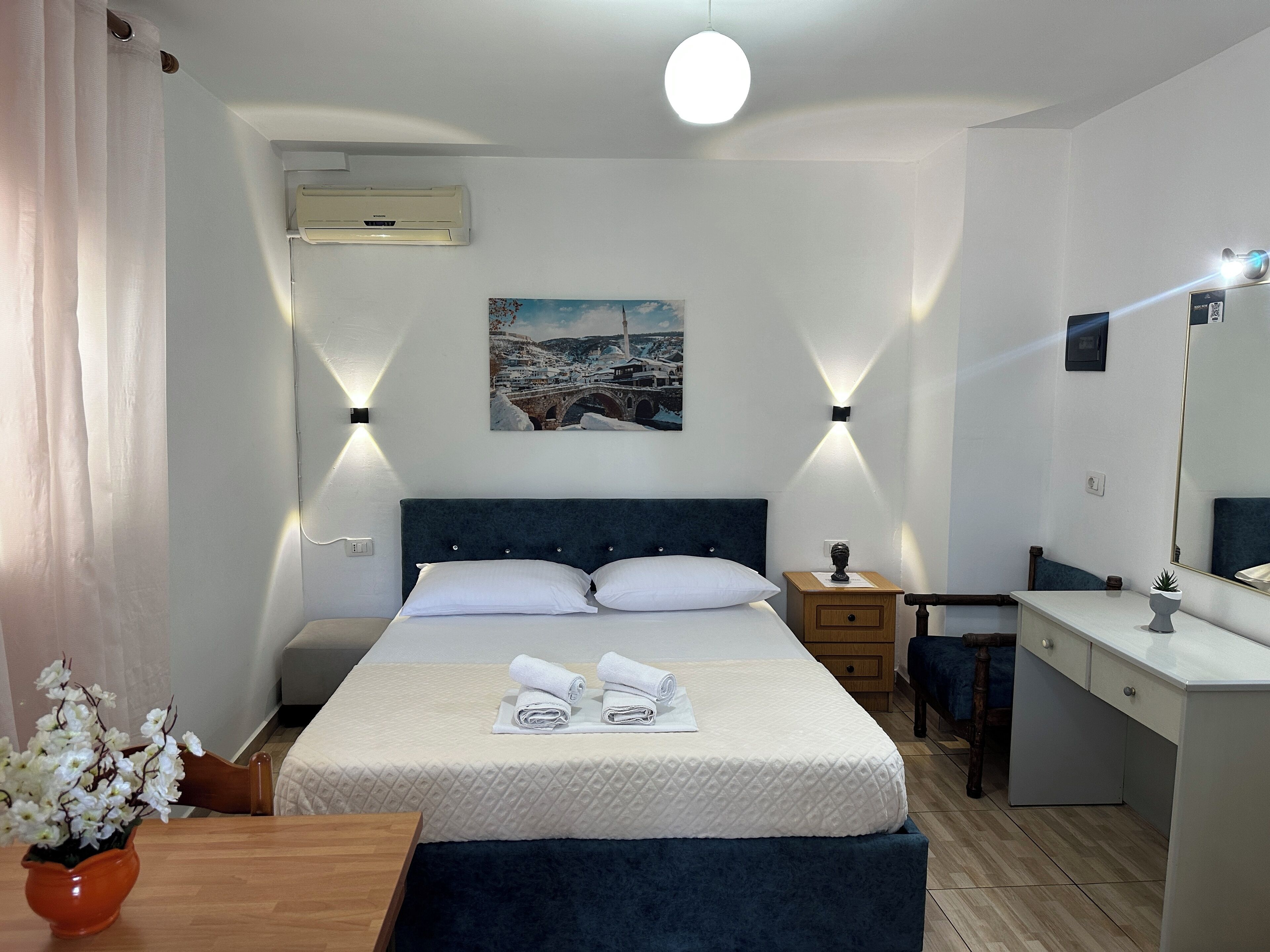 A hotel room with a bed, bedside table, nightstand, dresser, and a painting on the wall.