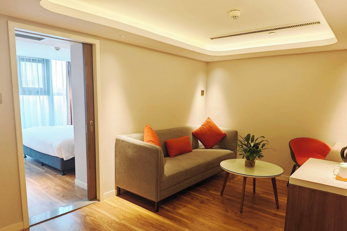 A modern hotel room with a wooden floor, a sofa with orange cushions, a small round table with a plant, and a bed in the adjacent room.