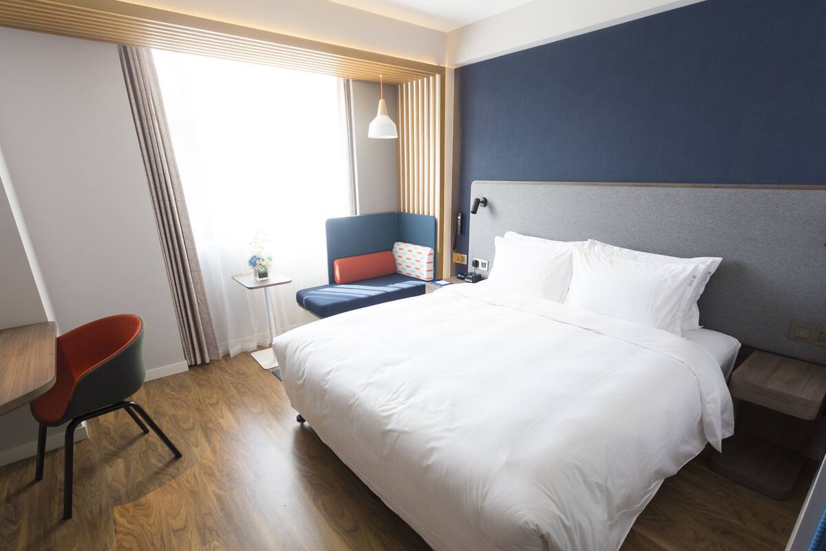 A hotel room with a large bed, a desk, a chair, and a window with curtains.