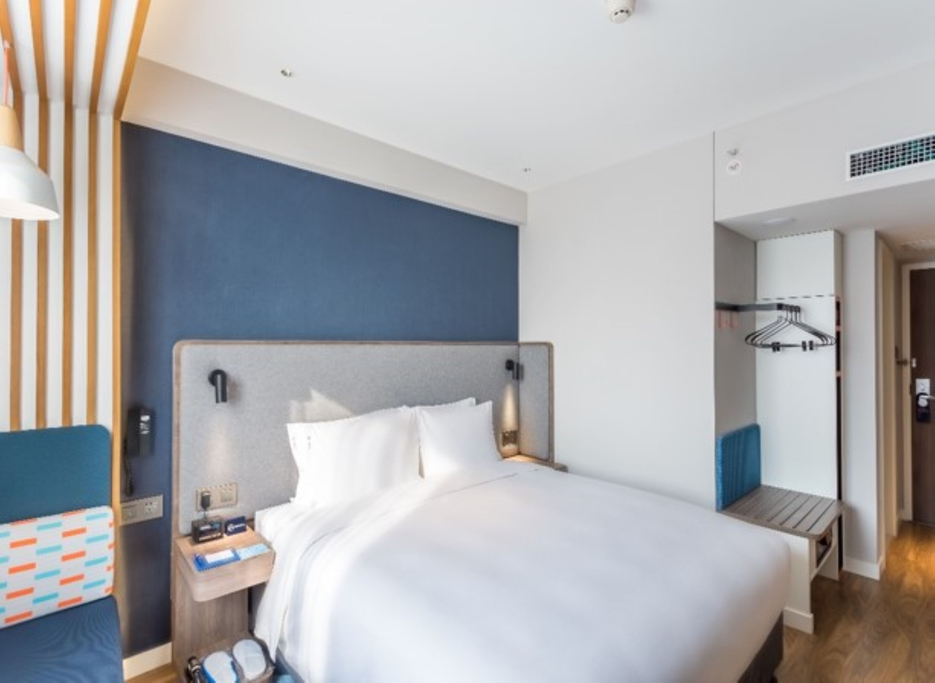 A hotel room with a bed, bedside table, lamp, and a view of a hallway with a coat rack.