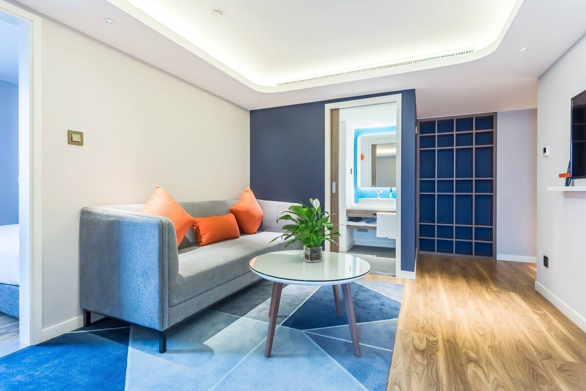 A modern living room with a grey sofa, orange cushions, a glass coffee table, and a potted plant.