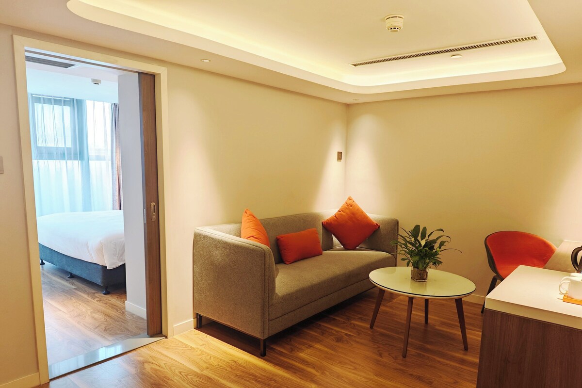 Ein modernes Hotelzimmer mit Holzboden, einer Couch mit orangen Kissen, einem kleinen runden Tisch mit einer Pflanze und einem Bett im angrenzenden Raum.