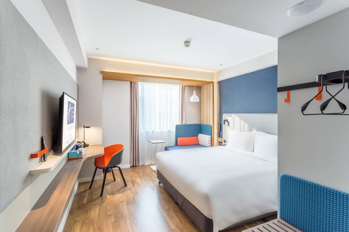 A modern hotel room with a large bed, a desk with a chair, a TV, and a window with curtains.
