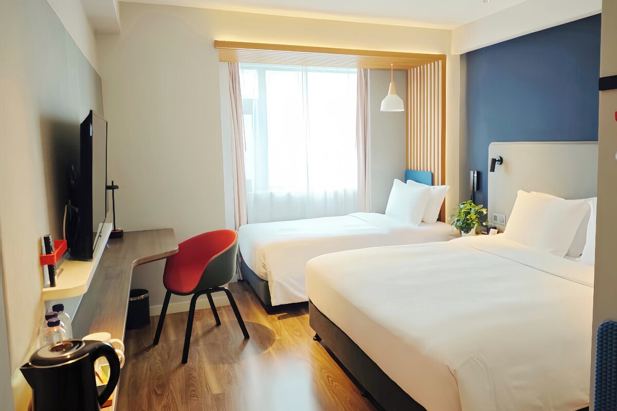 Ein Hotelzimmer mit zwei Betten, einem Schreibtisch mit Stuhl, einem Fernseher und einem Fenster mit Vorhängen.