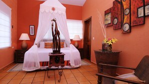 A bedroom with a canopy bed, a nightstand with a lamp, a chair, and a small table with a vase of flowers. - Bamboo Manor (Playa del Carmen)