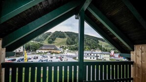 A view from a covered balcony with a wooden railing, overlooking a parking lot and buildings, including a hotel with a distinctive roof. - Steirer Apartment with Small Balcony - Steirer Apartment (Semmering-Kurort)