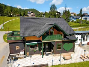 Un bâtiment en bois doté d’un balcon vert, d’un espace extérieur avec des sièges à motifs à carreaux et d’un motif en forme de cœur sur le côté.