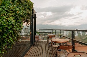 Una terraza de madera con mesas y sillas, rodeada de follaje y con vistas a un paisaje urbano.