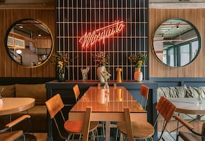 Un interior de restaurante con un letrero de neón que dice "Mosquito", paneles de madera, mesas redondas y sillas.