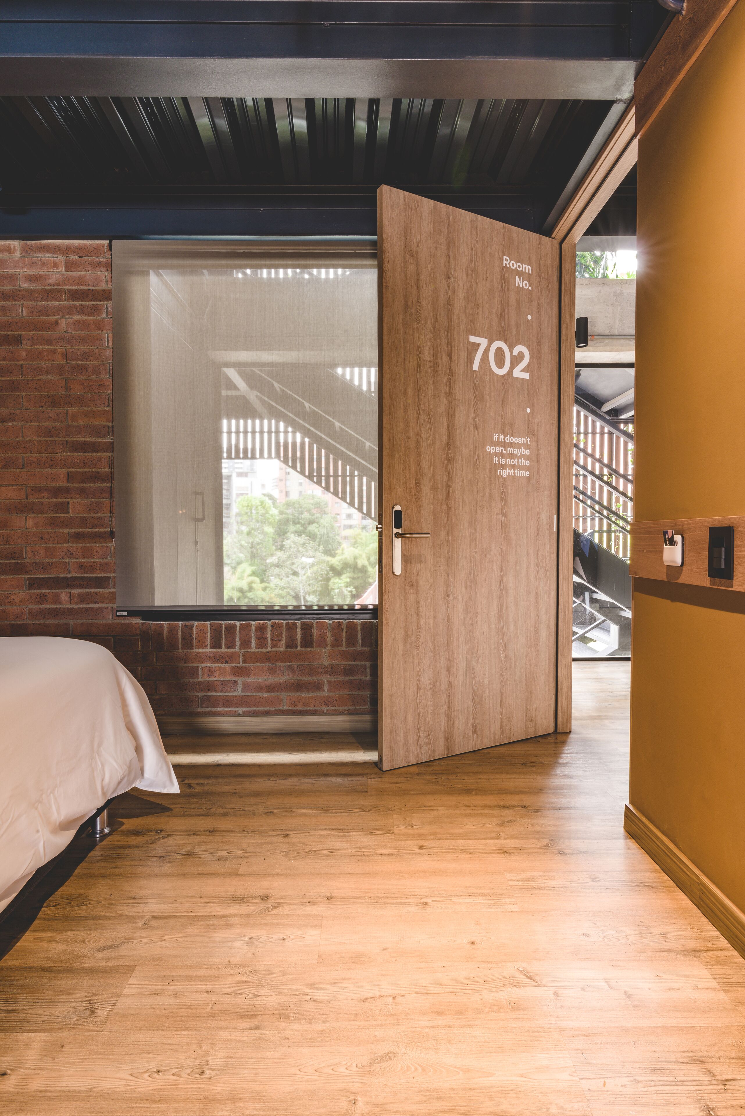A hotel room with a wooden door labeled "Room No. 702," a large window, and a view of a staircase and greenery outside.