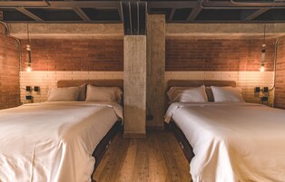 Habitación de hotel con dos camas, pared de ladrillo, tuberías del techo a la vista y lámparas colgantes.
