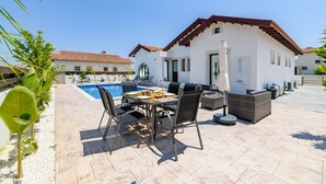 Un patio con mesa y sillas, una piscina y un edificio blanco con techo rojo.