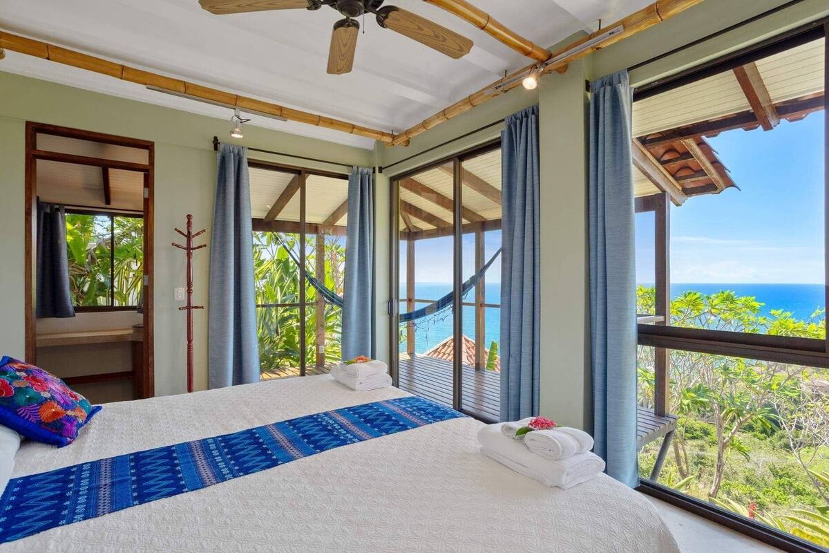 Een slaapkamer met een groot bed, een houten plafondventilator en uitzicht op de oceaan.