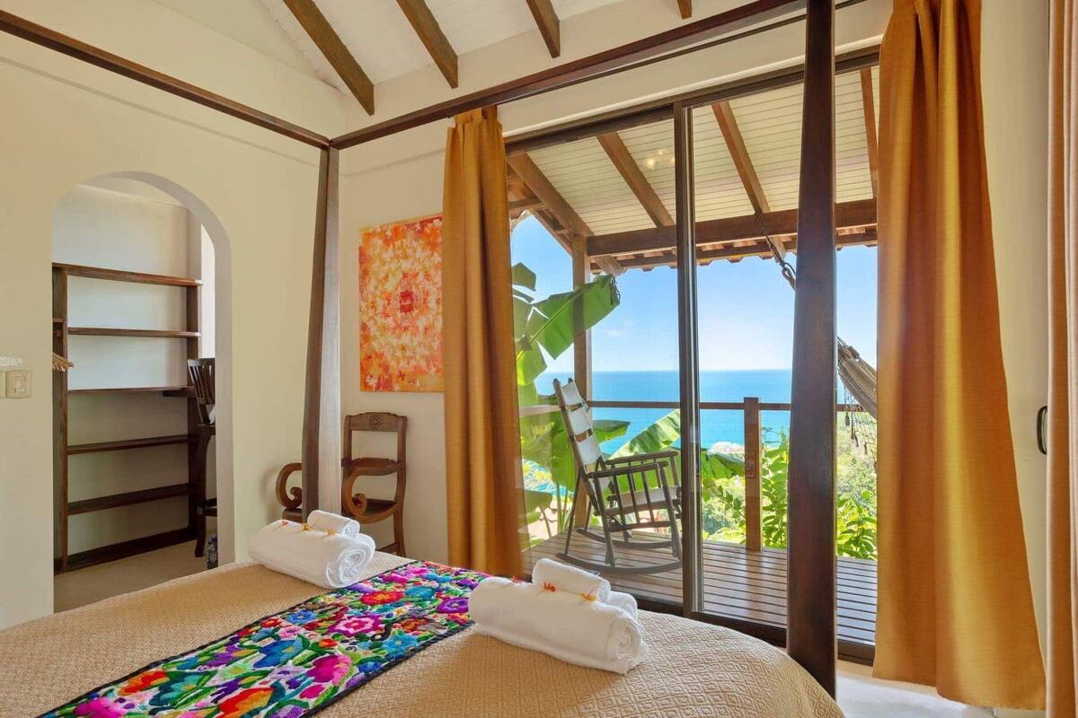 Een slaapkamer met een groot bed, uitzicht op de oceaan en een balkon met een stoel.