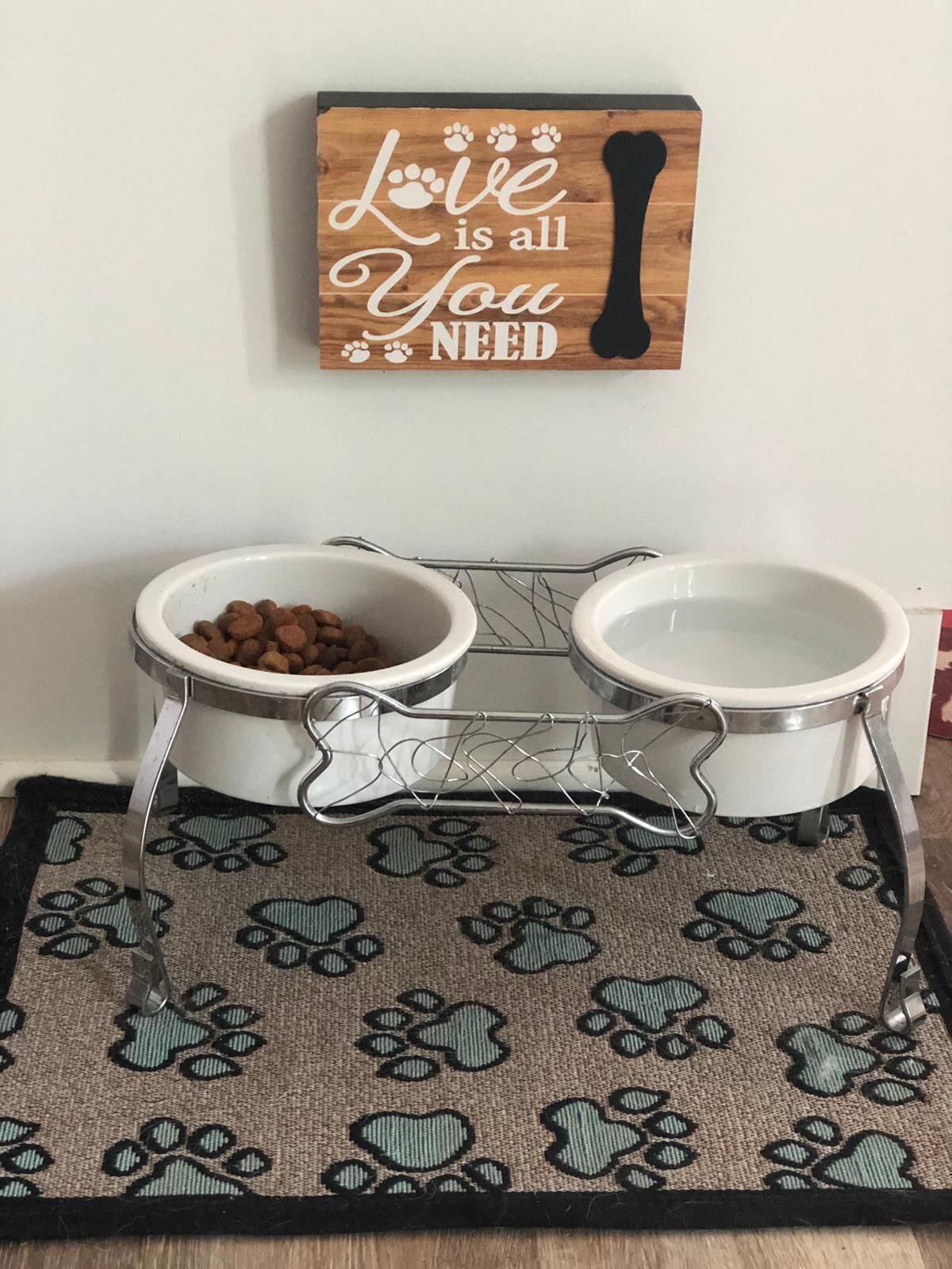 A pet feeding station with two bowls on a wire rack, a wooden sign above, and a patterned rug below.