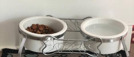 A pet feeding station with two bowls on a wire rack, a wooden sign above, and a patterned rug below.