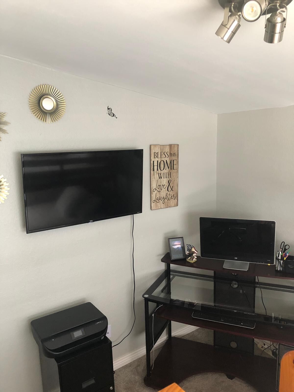 A home office with a large flat-screen TV mounted on the wall, a desk with a computer, and a trash bin.