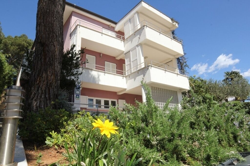 A multi-story residential building with balconies, surrounded by greenery and a large tree.