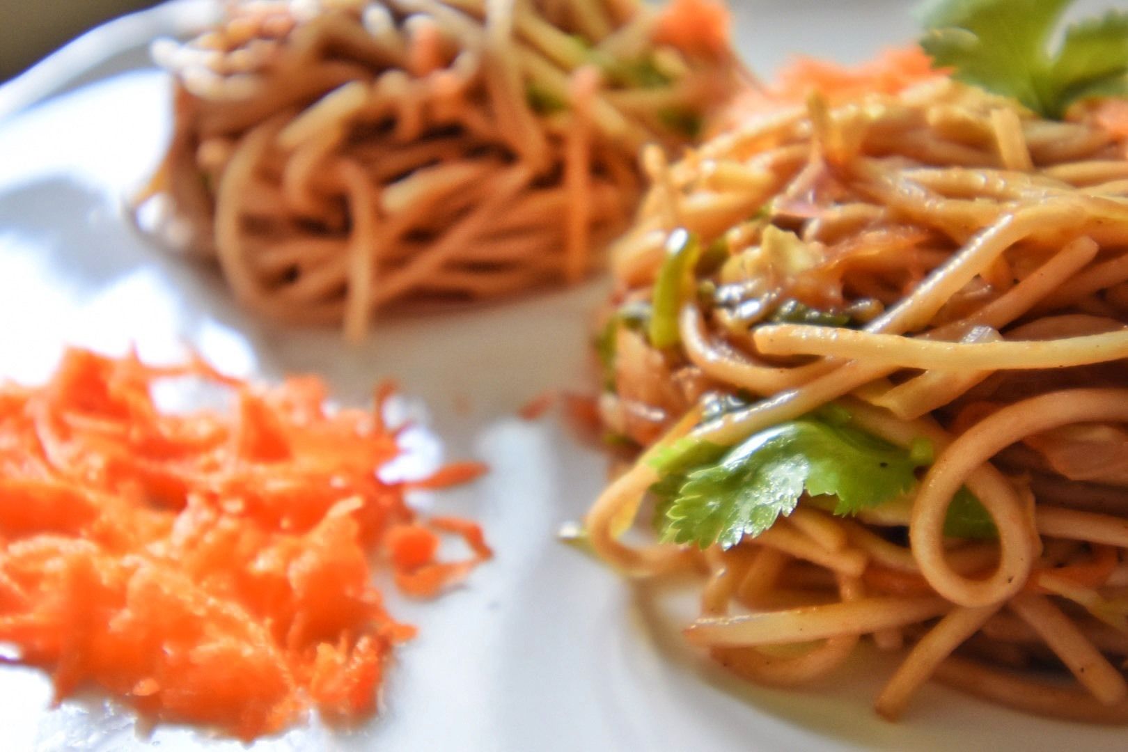 A plate of noodles with vegetables and a side of shredded carrots.