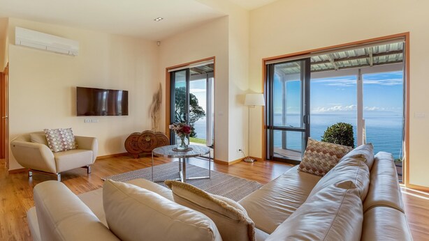 Un salon spacieux avec un grand canapé, un fauteuil, un téléviseur et une vue sur la mer grâce à de grandes fenêtres.