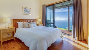 Une chambre à coucher avec un grand lit, une tête de lit en bois, des tables de chevet avec des lampes, un tableau encadré au mur et une vue sur la mer par une porte-fenêtre coulissante.
