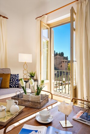 Une pièce bien éclairée avec un balcon, un canapé, une table sur laquelle se trouve un plateau avec des articles pour le thé, et une vue sur un bâtiment historique.