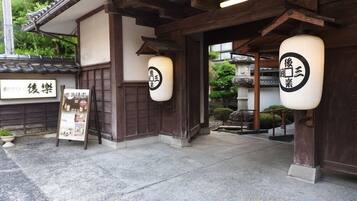 A traditional Japanese building with wooden beams and a tiled roof. There are two hanging paper lanterns with Japanese characters. A signboard with Japanese text is visible on the left.