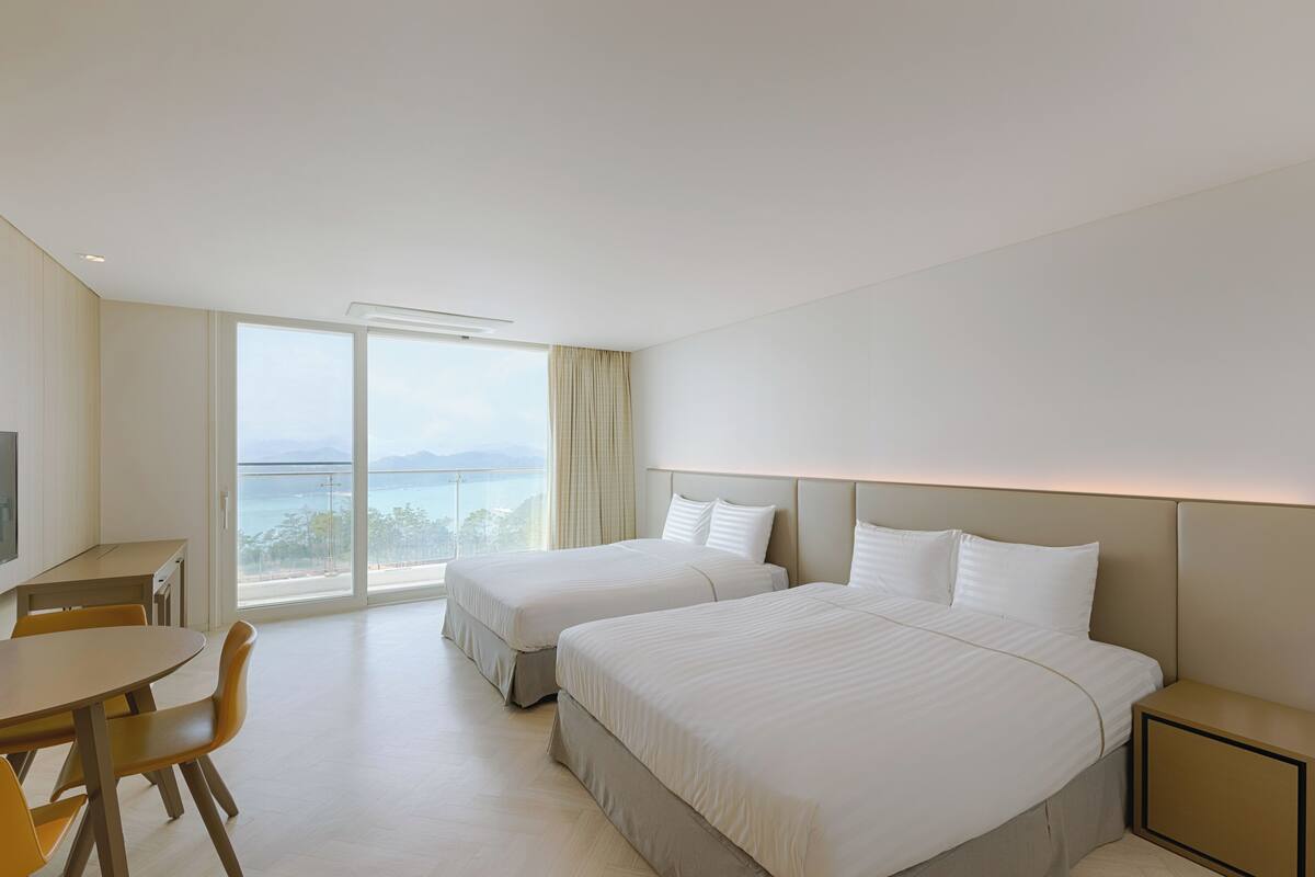 Habitación de hotel con dos camas, un escritorio y un amplio ventanal con vista al mar.