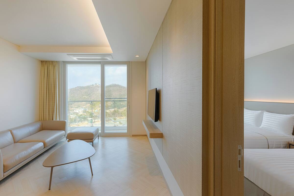 Una habitación de hotel moderna con un sofá, una mesita, un televisor montado en la pared y una cama con ropa blanca.