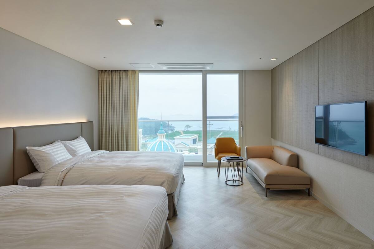 Habitación de hotel con dos camas, un sofá, un televisor y un ventanal con vista a la ciudad.