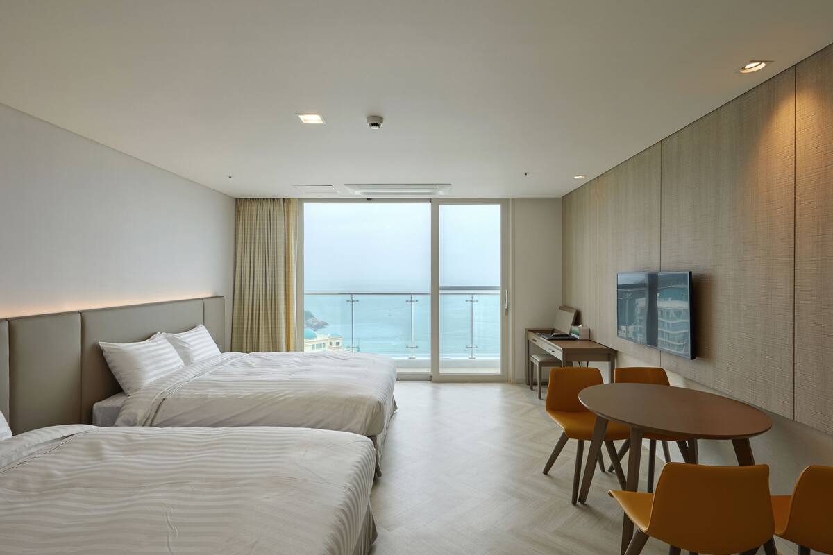 Habitación de hotel con dos camas, una mesa de comedor con sillas, televisión y un amplio ventanal con vista al mar.