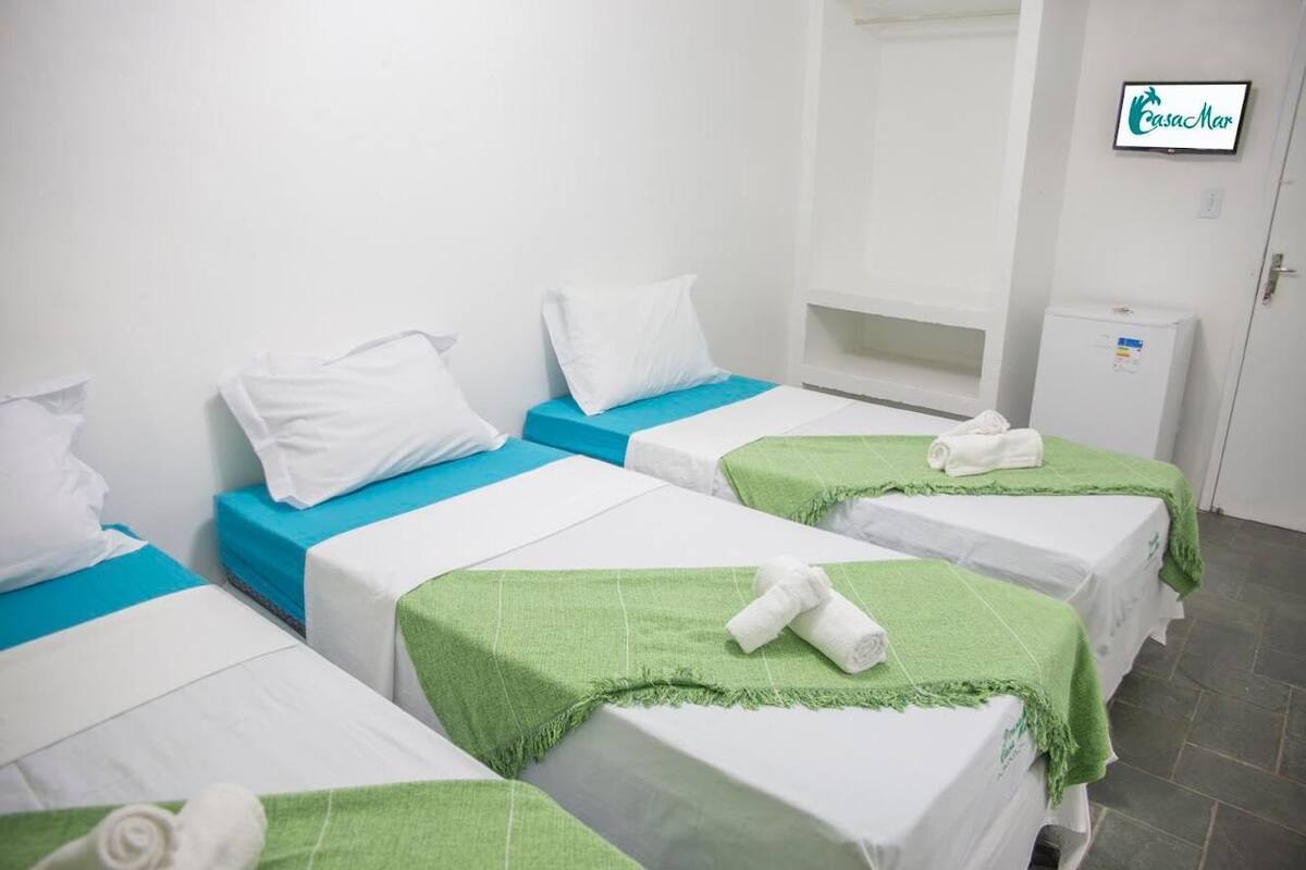 Ein Hotelzimmer mit vier Betten, jedes mit weißen Leinen und grünen oder blauen Akzenten. Ein Fernseher ist an der Wand montiert, und ein kleiner Schrank ist sichtbar.