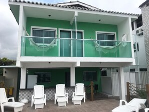 Un edificio de dos pisos con fachada verde y blanca, un balcón con sillas blancas y un patio con baldosas.