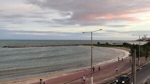 A coastal road with a paved walkway alongside a beach, a parked car, and a distant city skyline. - CasaMar Pousada (Paulista)