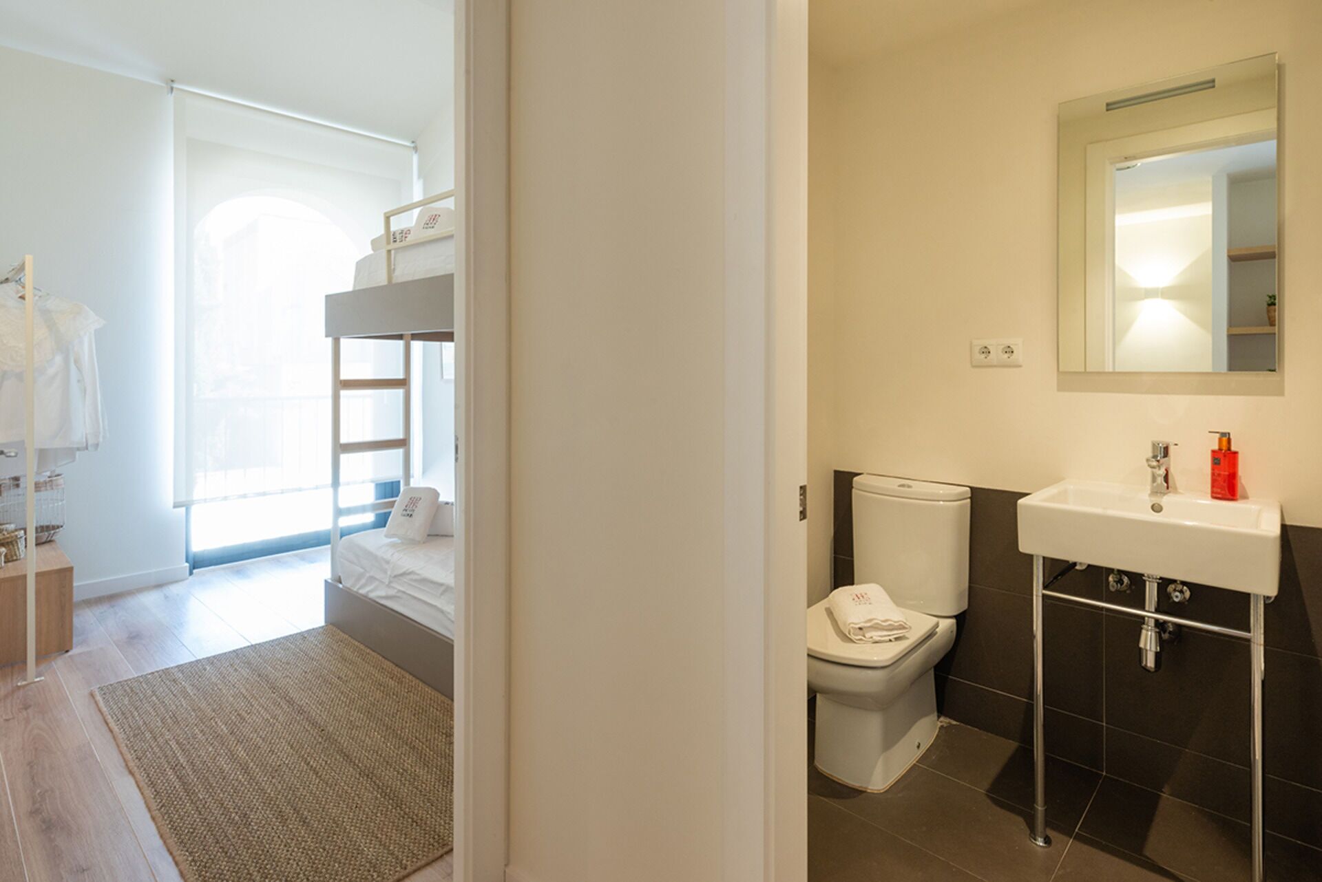 A bedroom with a bunk bed, a small bathroom with a toilet and sink, and a view of the outside.
