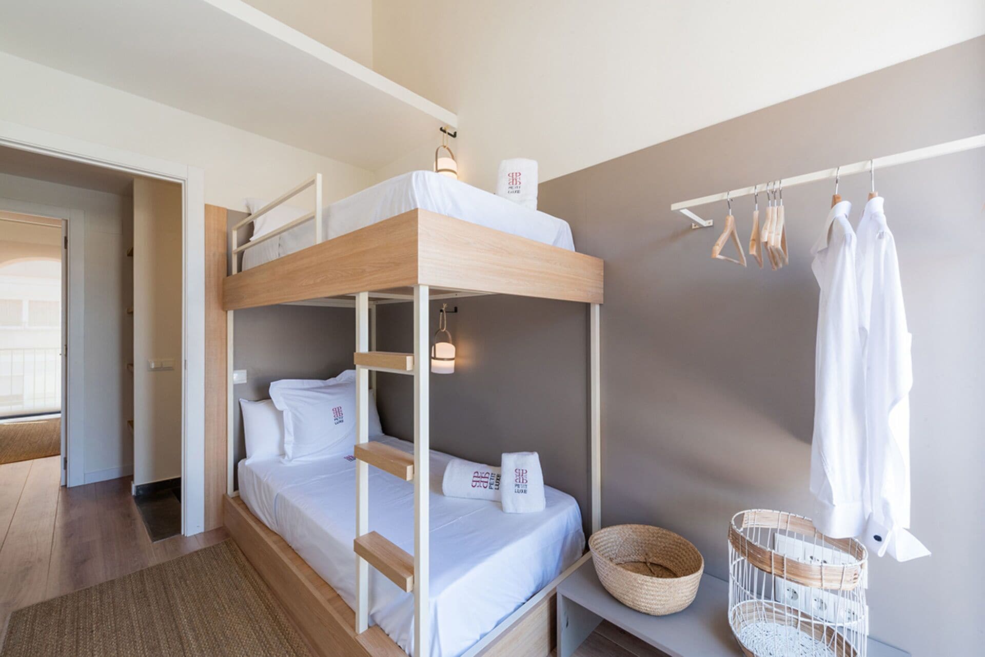 Een slaapkamer met een stapelbed met houten frame, witte beddengoed, een klein nachtkastje met een mand en een kledingrek met hangende kleding.