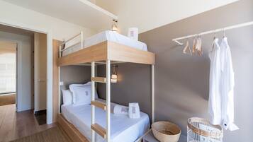 Een slaapkamer met een stapelbed met houten frame, witte beddengoed, een klein nachtkastje met een mand en een kledingrek met hangende kleding.