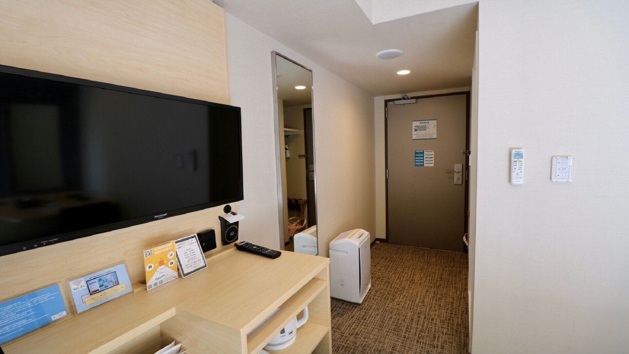 A hotel room with a flat-screen TV, a desk with a remote control, a small heater, and a door with a sign.