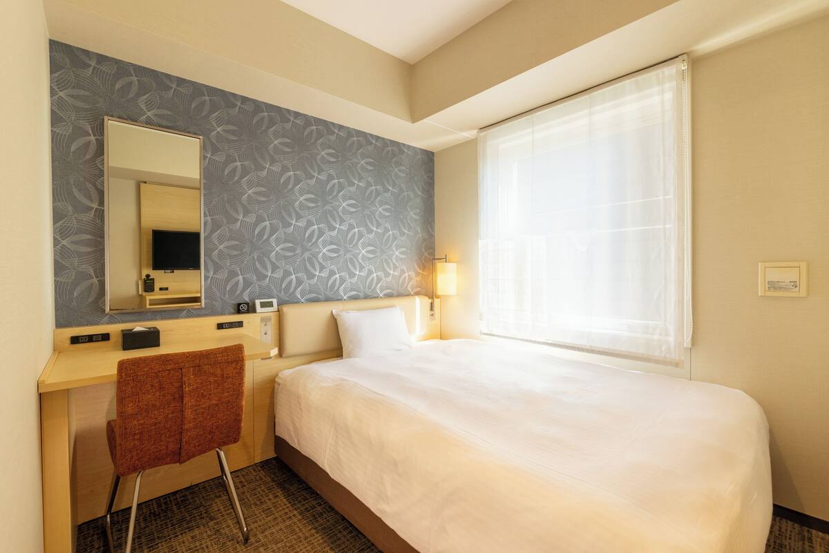 Habitación de hotel con cama, escritorio, silla, espejo, televisor y ventana con cortinas.