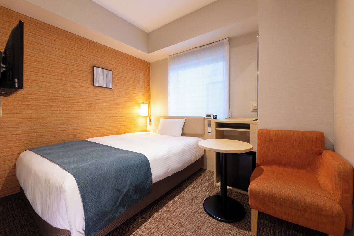 Habitación de hotel con cama, una silla, una mesita, televisor y ventana con cortinas.