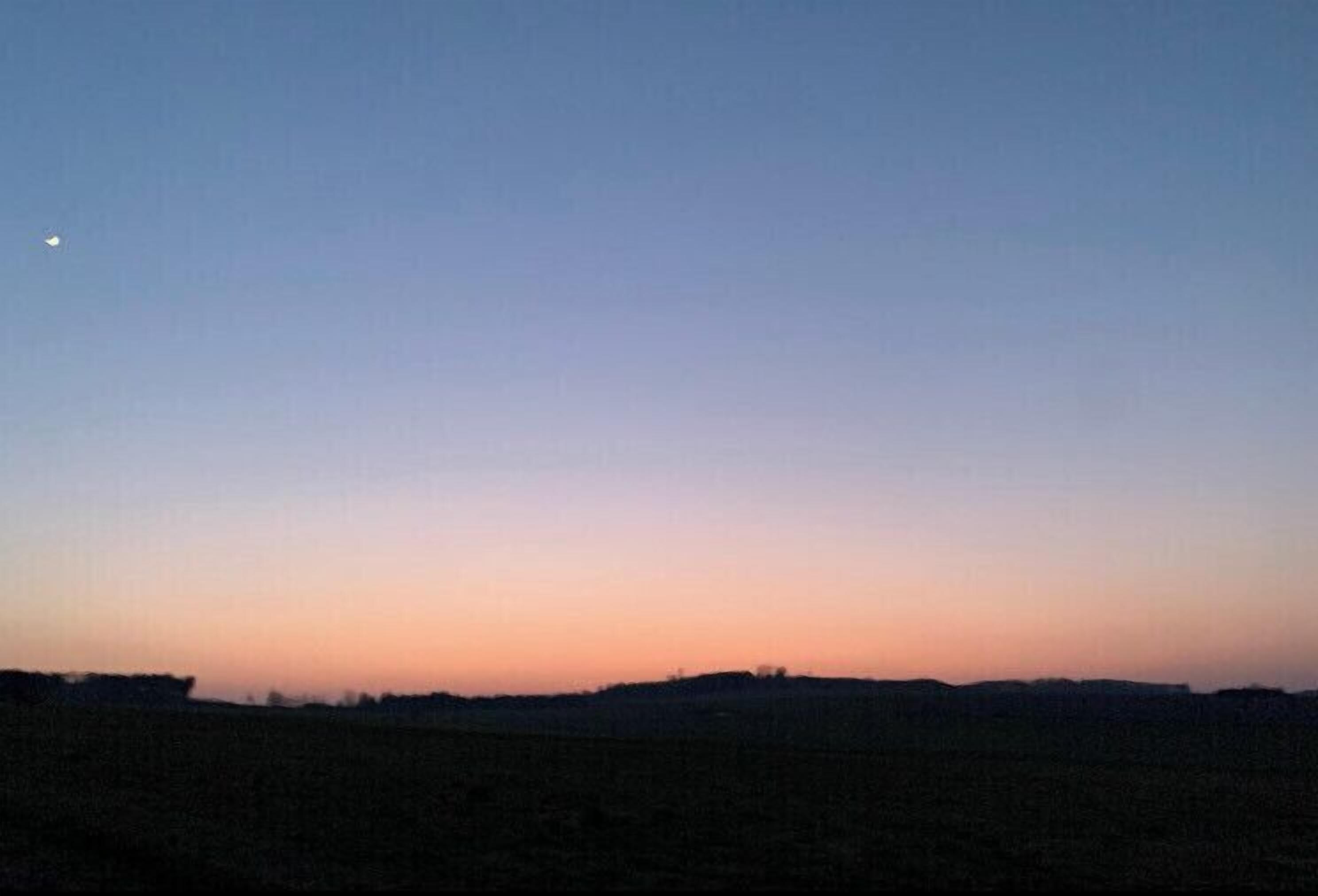 Eine flache Landschaft mit blauem Himmel und einem kleinen Mond in der oberen linken Ecke.