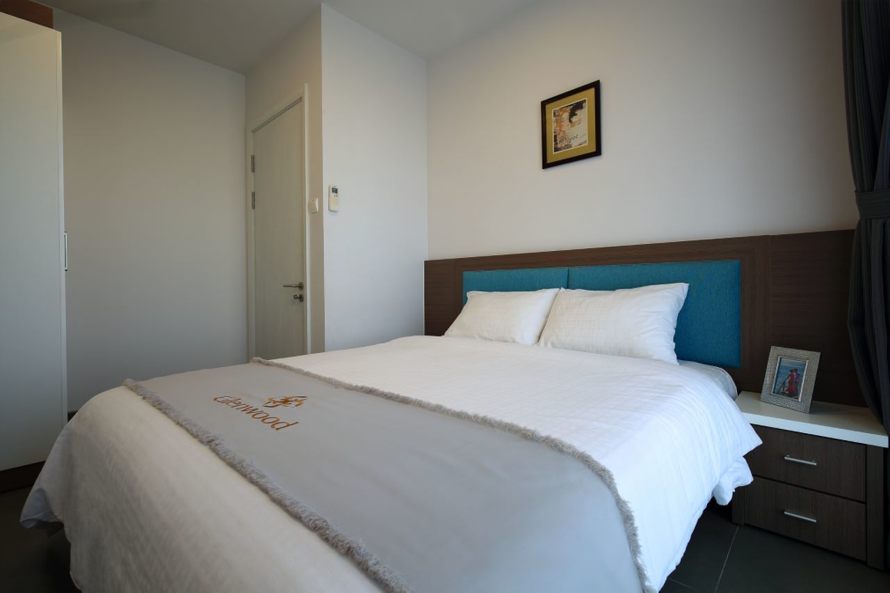 A neatly made bed with a white comforter and a decorative blanket, a wooden headboard, a bedside table with a picture frame, and a door to the left.