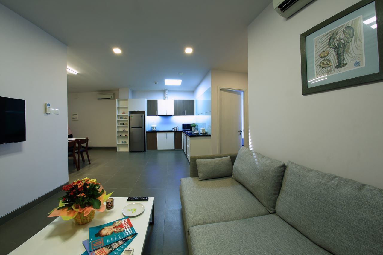 A modern living room with a grey sofa, a coffee table with magazines, and a TV on the wall. A kitchen is visible in the background with white cabinets and a refrigerator.