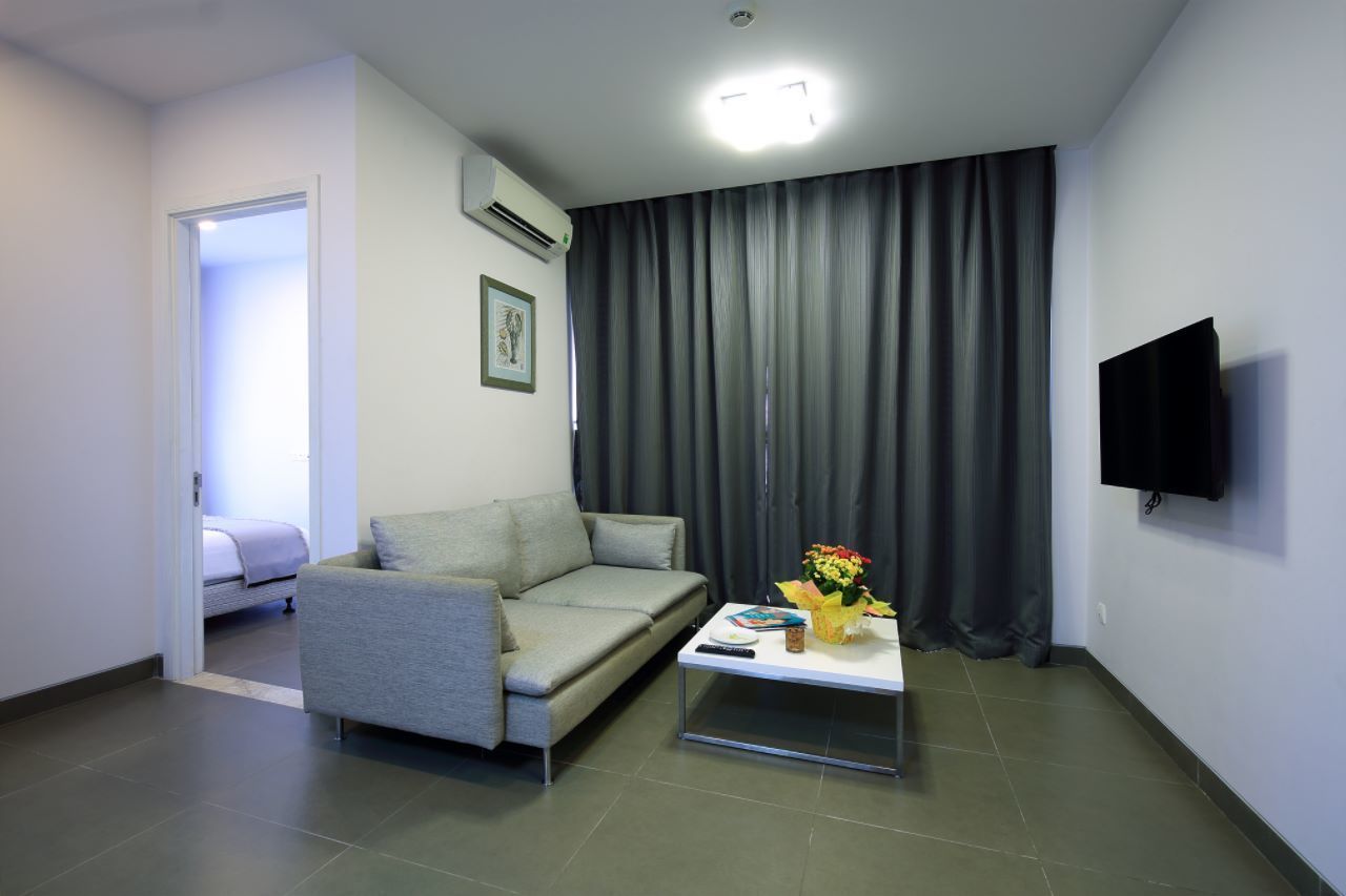 A modern living room with a grey sofa, a coffee table with a vase of flowers, a flat-screen TV mounted on the wall, and a large window with grey curtains.