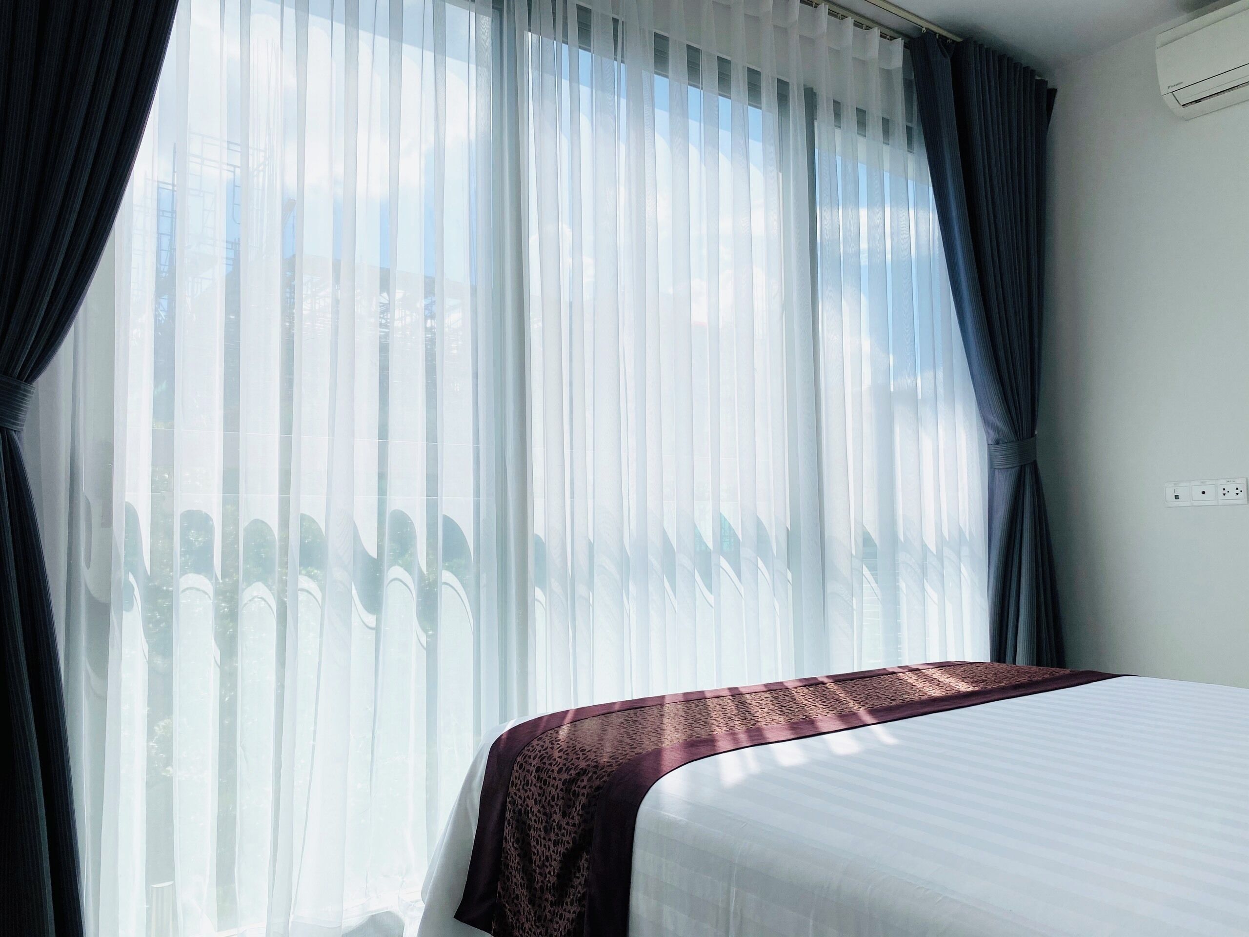A hotel room with a bed, sheer curtains, and an air conditioning unit.
