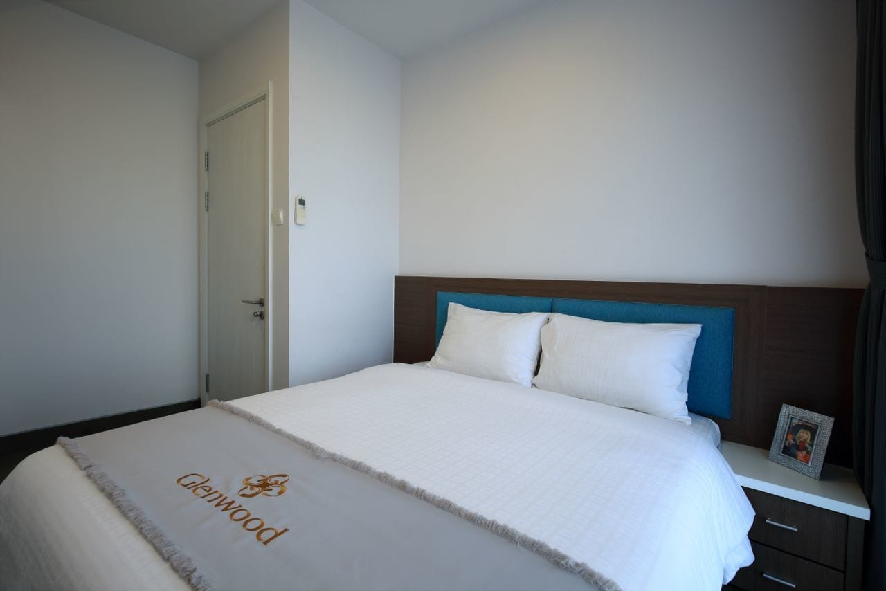 A neatly made bed with a white duvet and a logo on the bedspread, a wooden headboard, a bedside table with a picture frame, and a closed door.